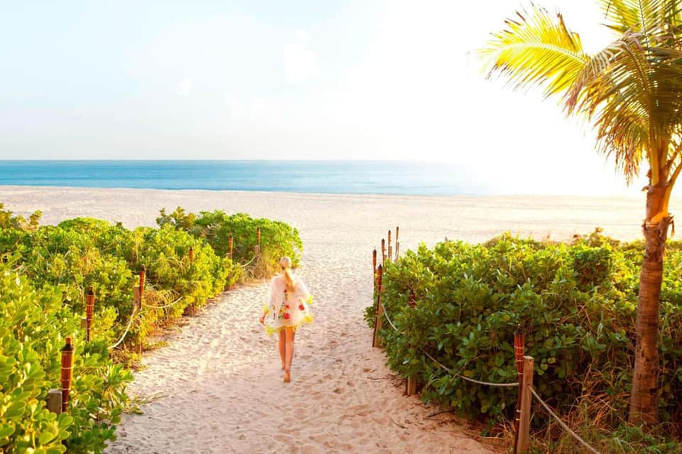 Woman in yellow dress walking on sandy beach path through dune vegetation toward ocean