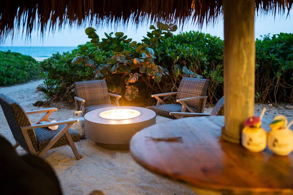 Beach fire pit lounge with woven chairs under thatched roof at sunset with ocean beyond