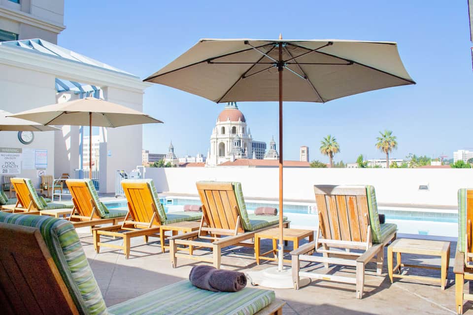 Rooftop pool deck with yellow lounge chairs, umbrellas, and city skyline view including a domed cathedral