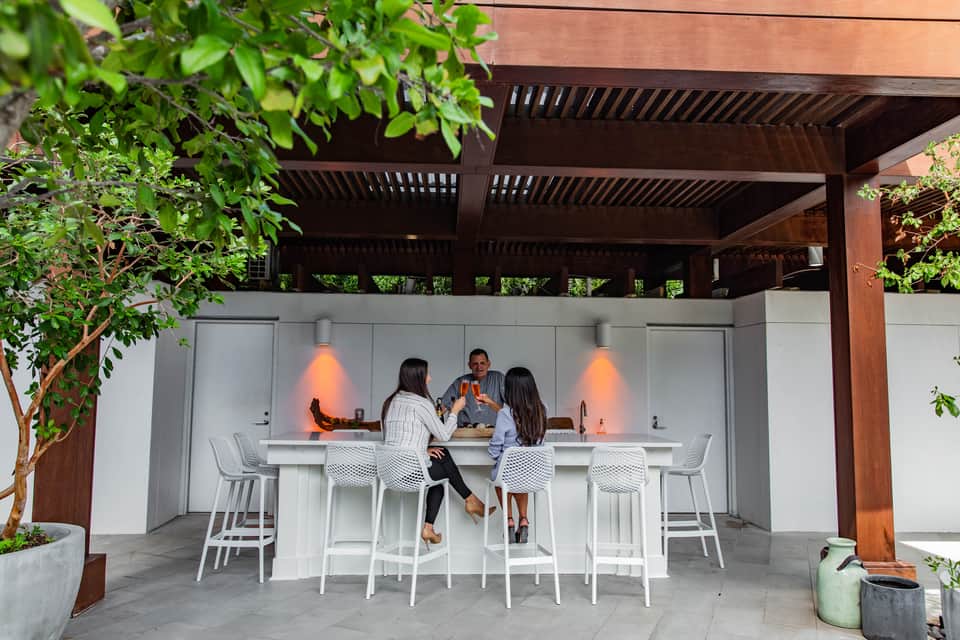 Outdoor bar counter with three guests, dark wooden pergola, and green foliage overhead