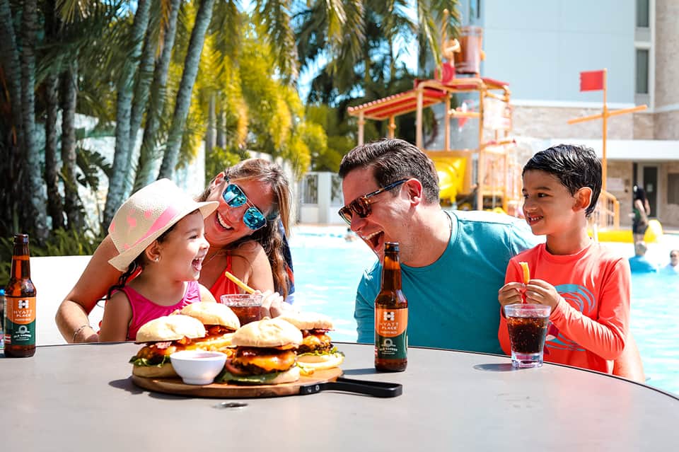 Family enjoying burgers and drinks poolside with colorful playground equipment visible in background