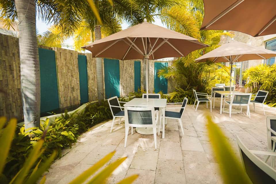 Shaded outdoor patio with white umbrellas, palm trees, and dining tables in a resort setting