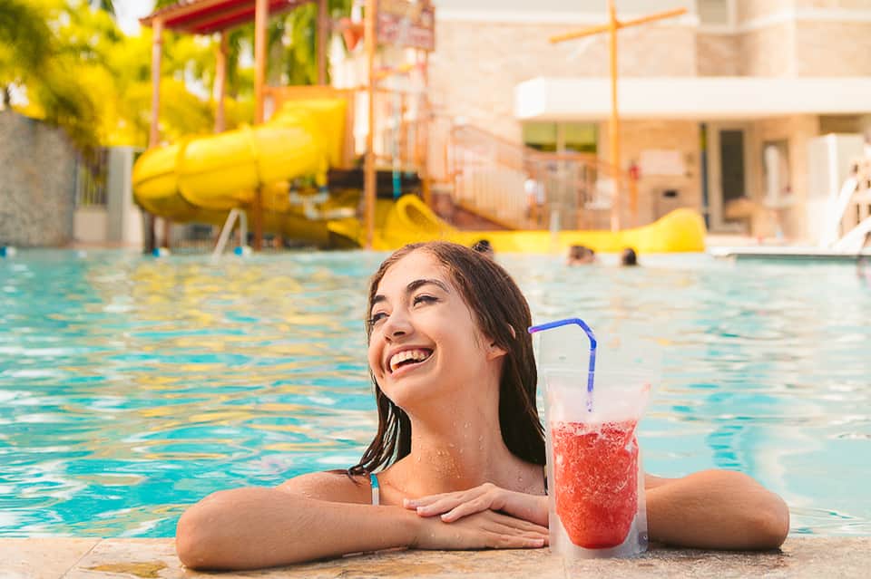 Smiling woman in pool holding red beverage with yellow water slide visible in background resort