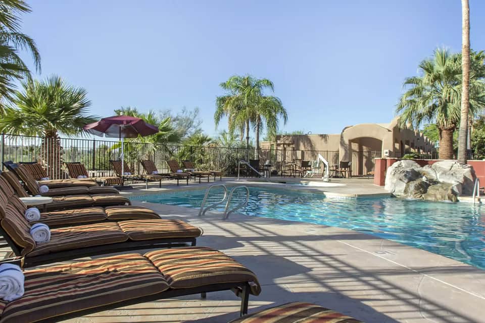 Resort pool with lounge chairs, red umbrella, palm trees, and architectural water feature