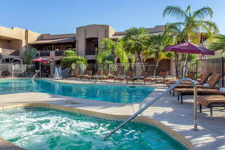 Resort pool with spa tub in foreground, lounge chairs, purple umbrellas, and desert landscaping