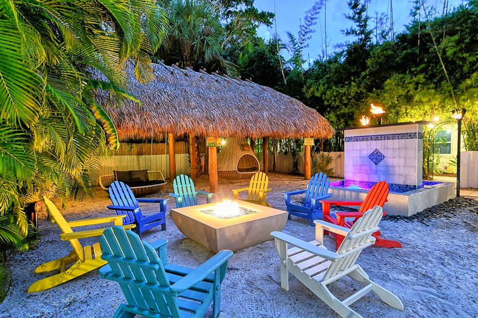 Illuminated beach cabana with thatched roof, colorful Adirondack chairs, and fire pit at night