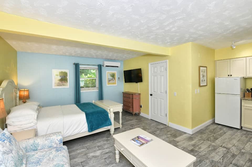 Bright hotel room with yellow walls, queen bed, kitchenette, and blue window treatments