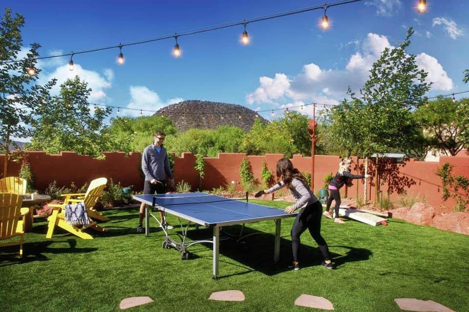 People playing table tennis on outdoor court with artificial grass, red walls, mountains, and string lights overhead