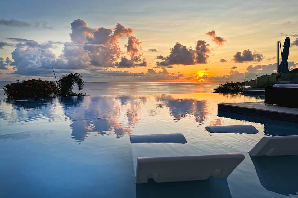 Stunning sunset view over an infinity pool blending into the ocean horizon with golden sky and fluffy clouds.