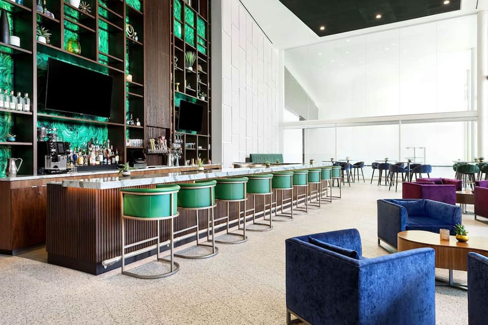 Modern hotel bar with green shelving, teal barstools, and lounge seating with city views