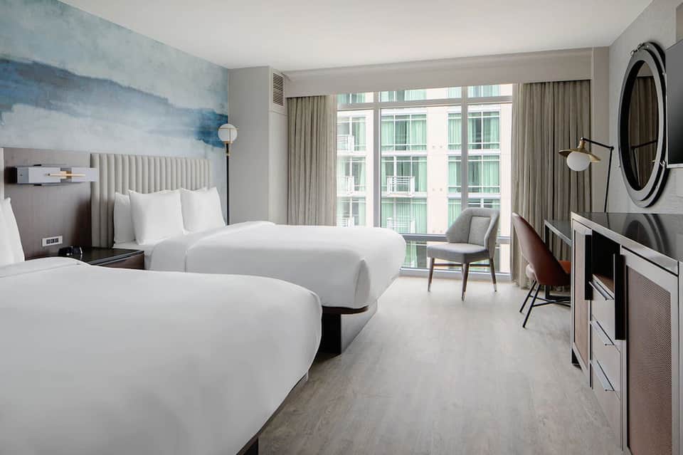 Contemporary hotel bedroom with two queen beds, blue accent wall art, floor-to-ceiling windows, and modern furnishings