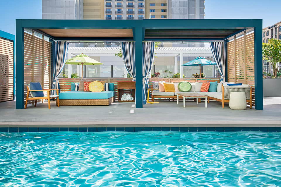 Resort poolside lounge area with turquoise pergolas, wood seating, and colorful cushions
