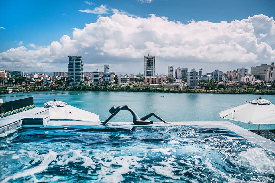 Rooftop pool with cityscape view of high-rise buildings reflected in turquoise water with white umbrellas