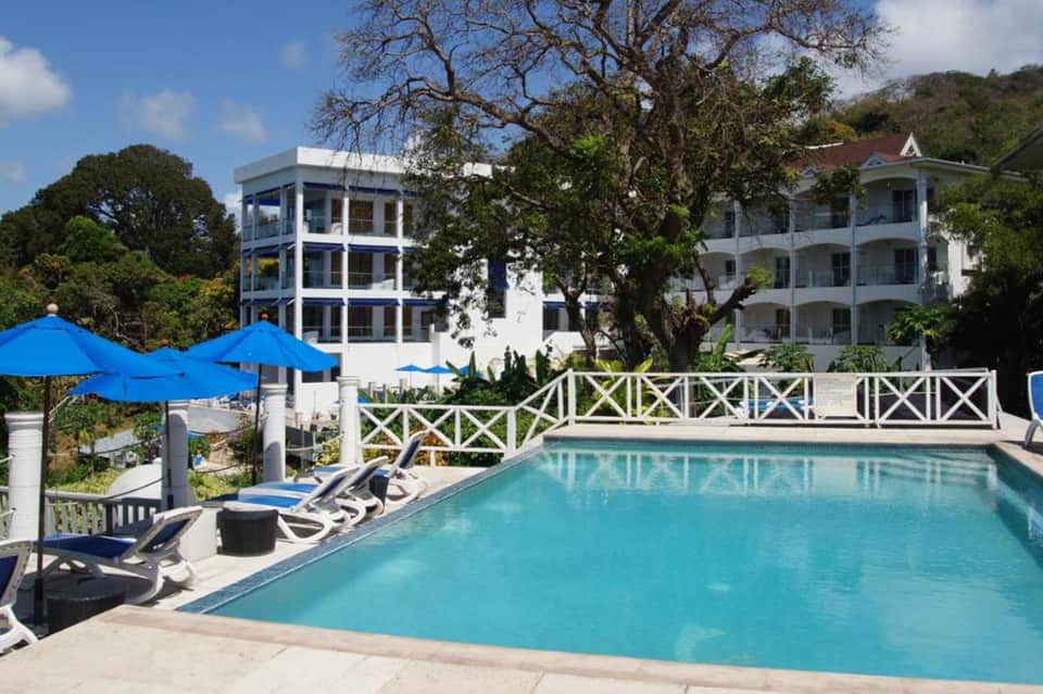 Resort pool with blue umbrellas, white colonial buildings, lounge chairs, and surrounding mature trees