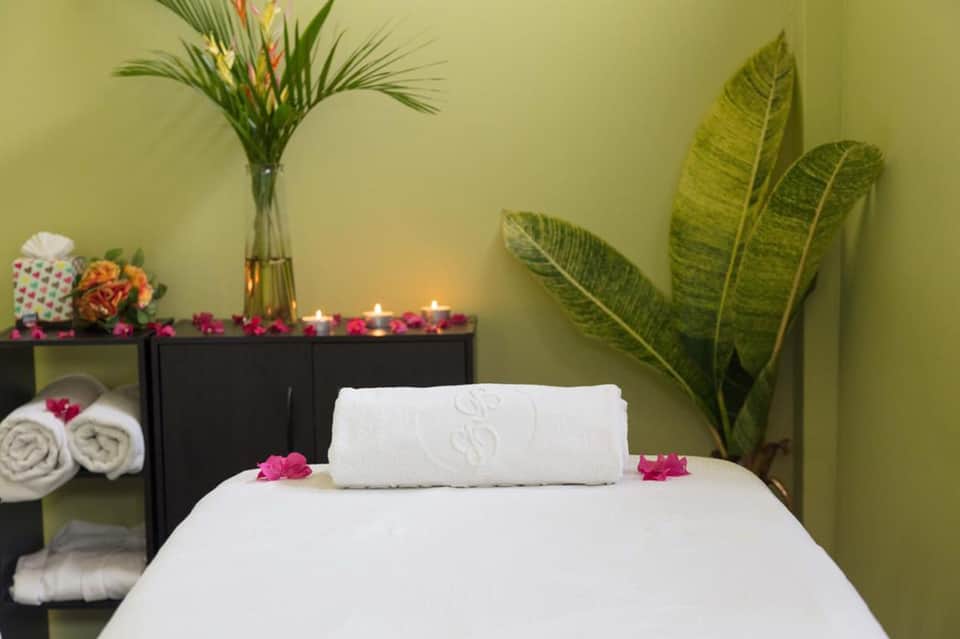 Spa treatment room with white massage bed, green walls, tropical plants, flowers, and candles