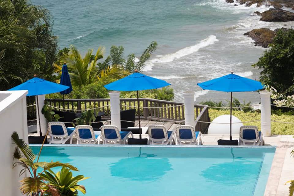 Oceanfront pool with blue umbrellas, lounge chairs, and ocean waves with rocky coastline