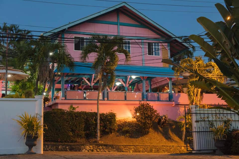 Pink and blue tropical house at dusk with palm trees and illuminated beachfront patio