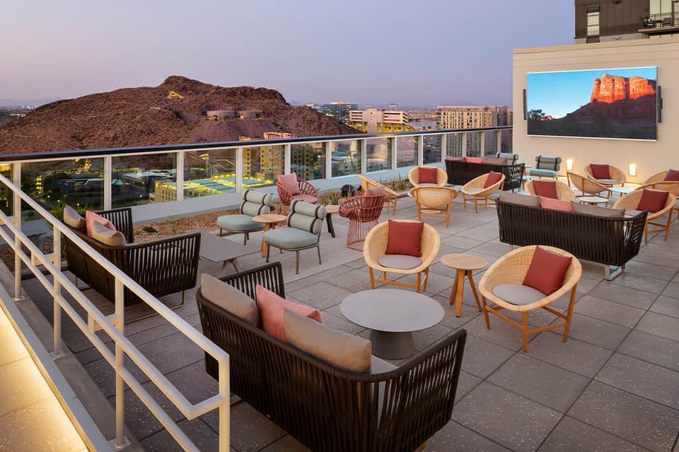Upscale desert rooftop lounge with woven chairs, fire features, mountain views, and outdoor screen