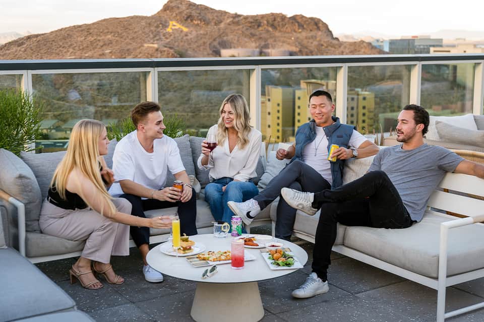 Five guests socializing on rooftop lounge with mountain views, appetizers, and drinks