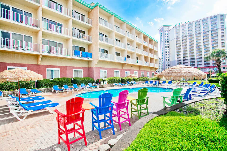 Resort pool area with colorful Adirondack chairs, thatched umbrellas, and multi-story beachfront hotel building