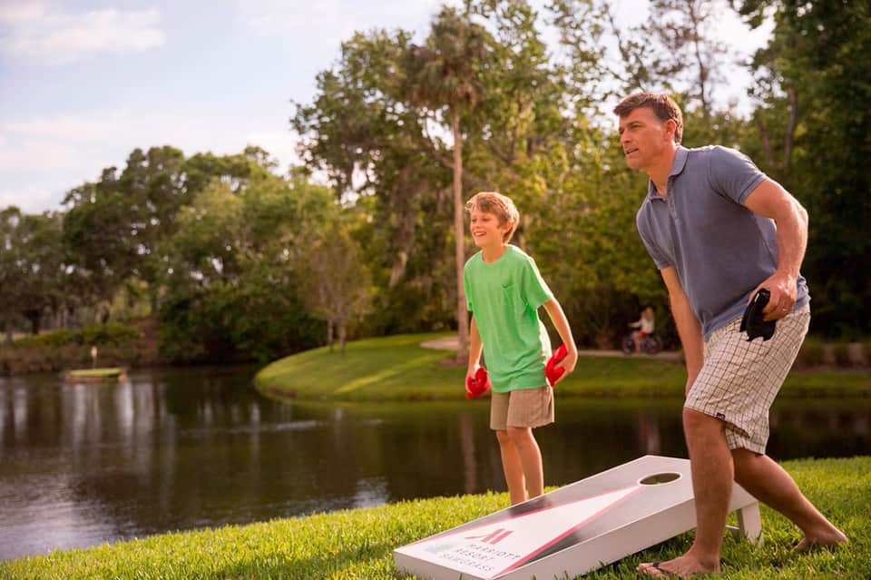 Father and son playing cornhole by a pond surrounded by green lawn and trees