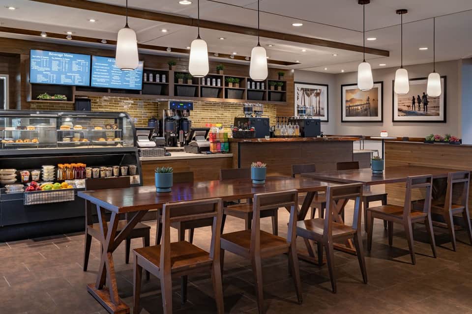 Contemporary hotel breakfast area with wooden tables, pendant lighting, bakery case, and menu board