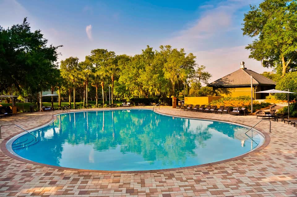 Large oval pool surrounded by brick deck, lounge chairs, yellow pavilion, and tall trees