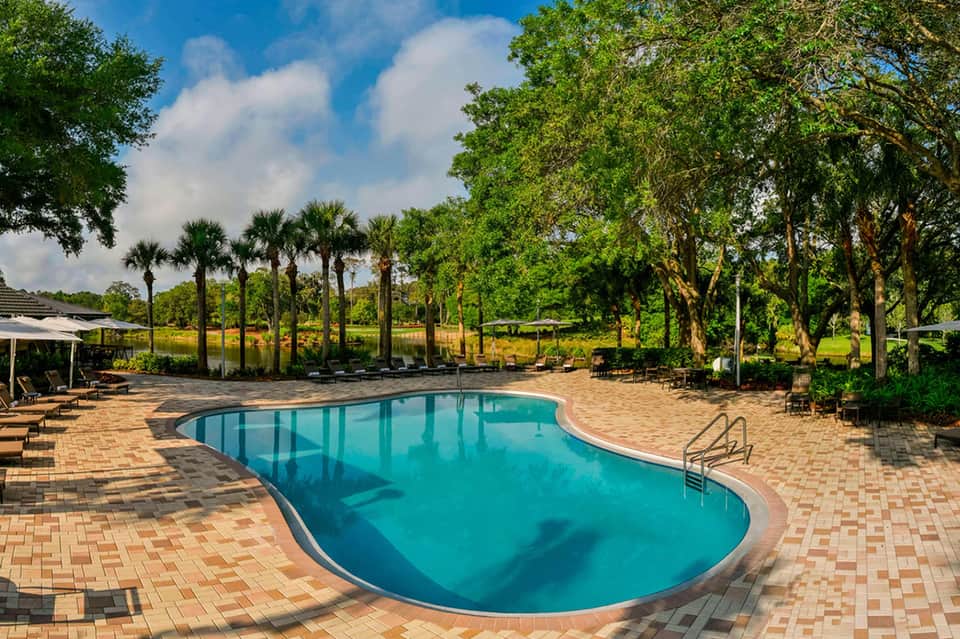 Resort pool surrounded by brick patio, mature oak trees, lounge chairs, and manicured grounds