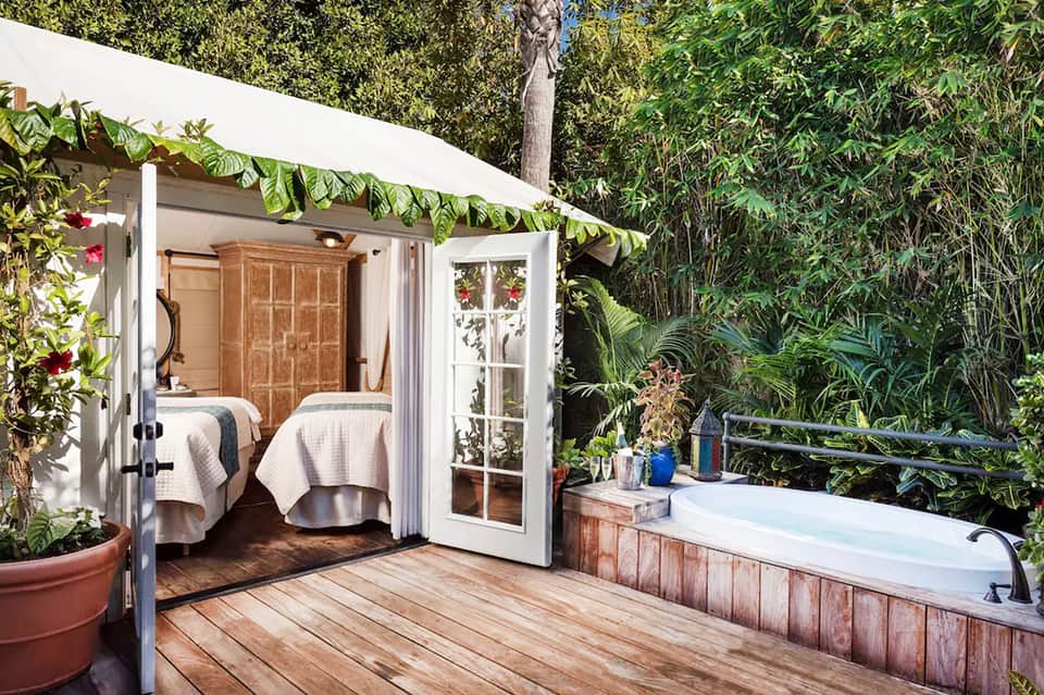 Tropical resort cabana with white frame, two beds, outdoor tub, and lush green vegetation