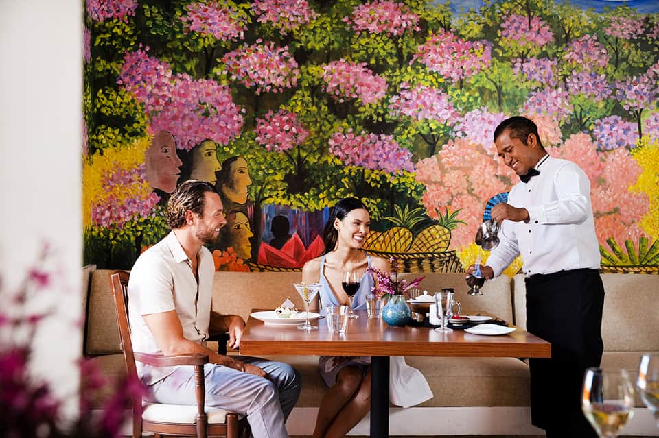 Fine dining restaurant with colorful floral artwork backdrop, server pouring wine for couple at table.