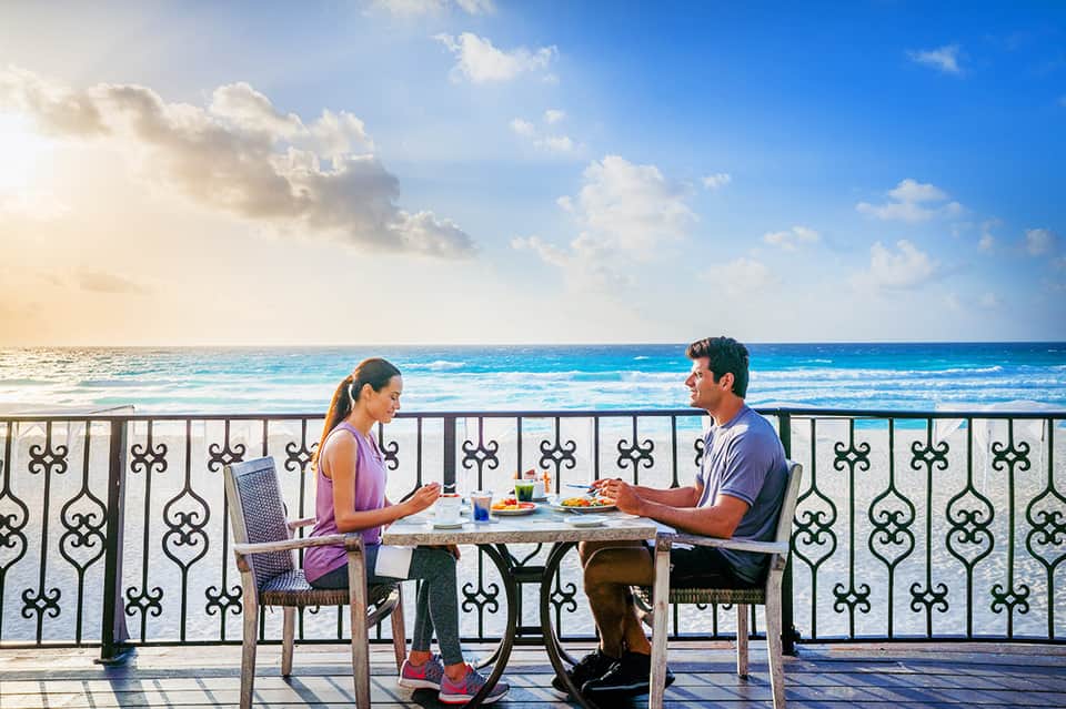 Couple dining at oceanfront table on balcony overlooking turquoise water and beach