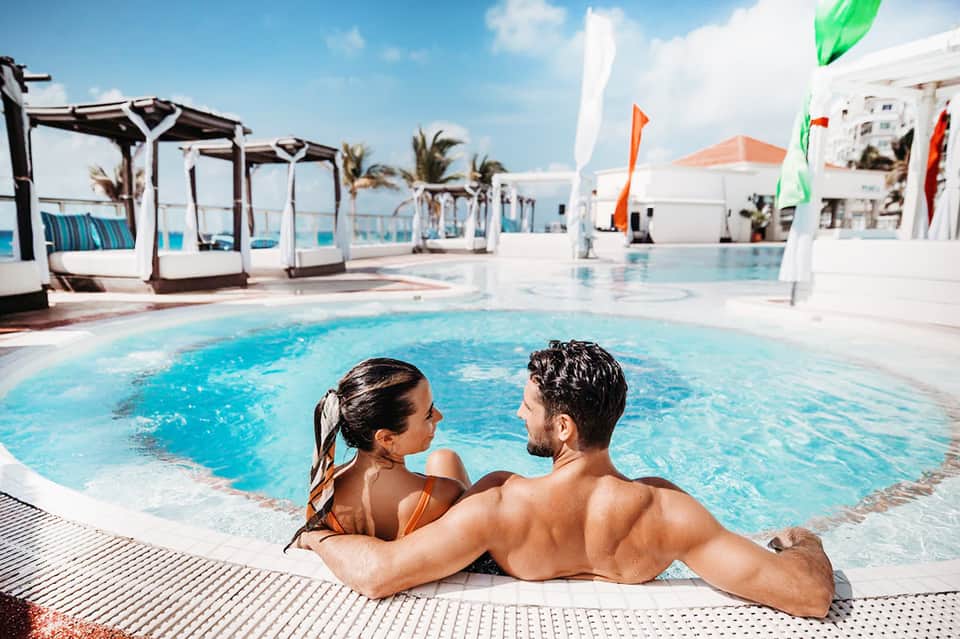 Resort pool with couple in water, cabanas, colorful flags, white lounge beds, and palm tree landscaping