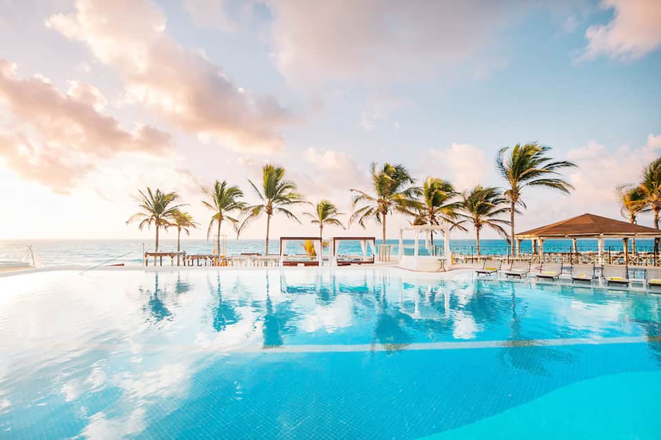 Oceanfront infinity pool with palm trees, white cabanas, and reflections under soft cloudy sky