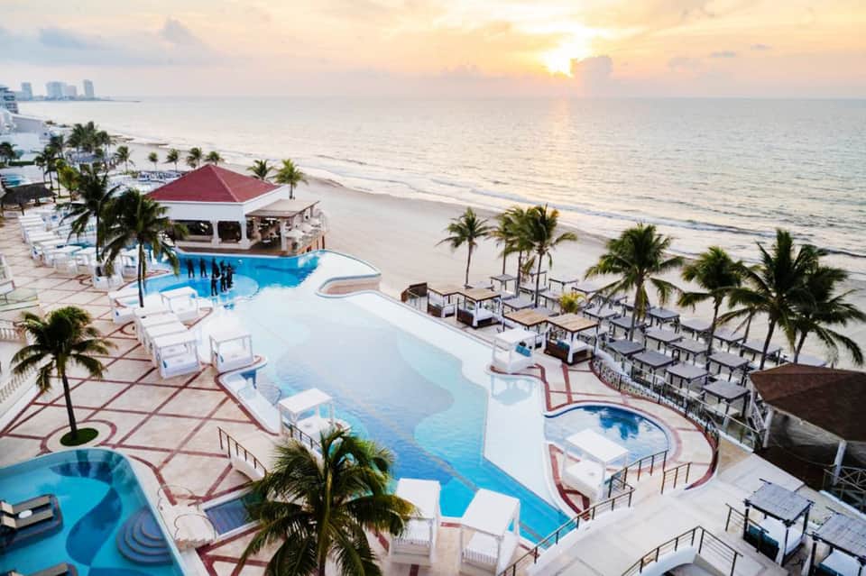 Aerial view of beachfront resort with multiple pools, palm trees, and ocean at sunset