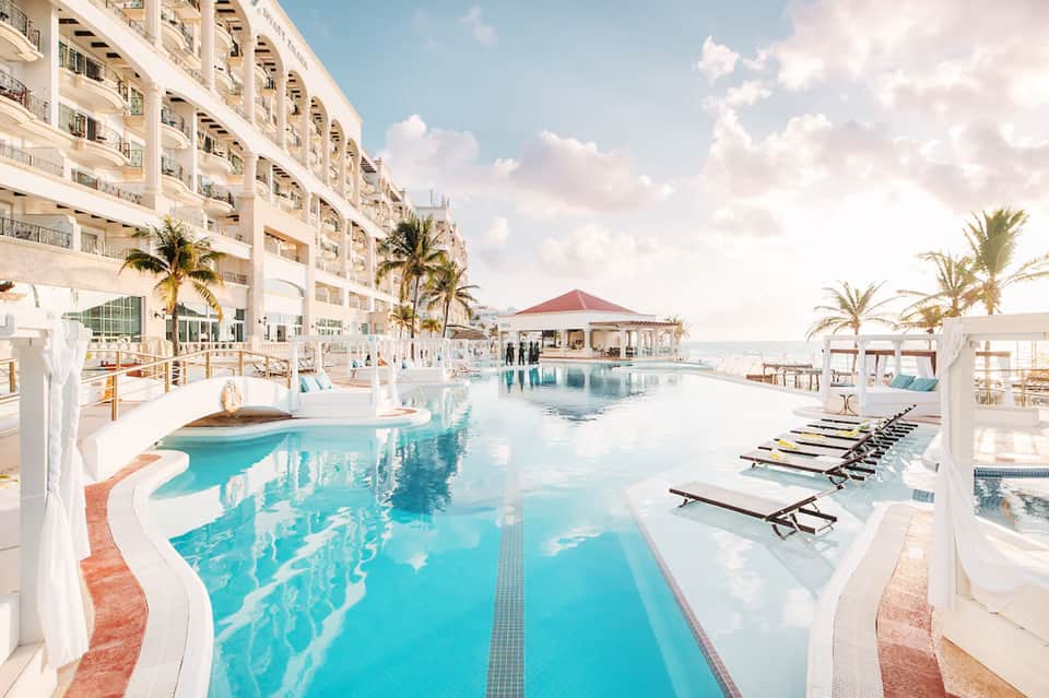 Curved resort pool with loungers, palm trees, beachfront building, umbrella shade structures, and ocean view