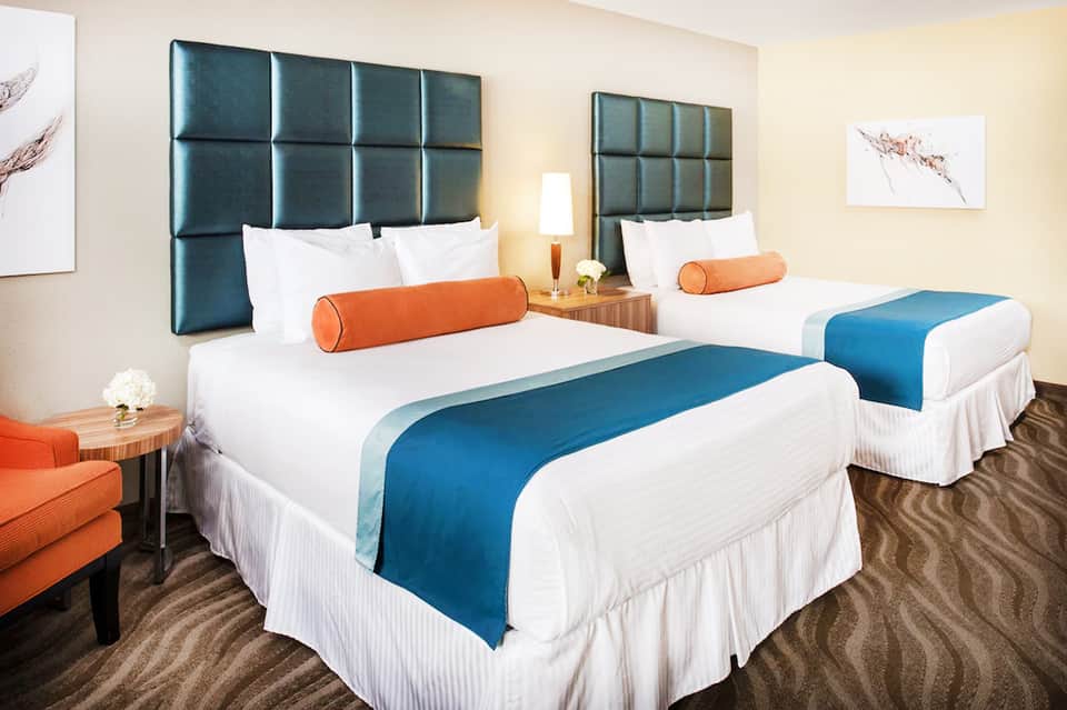Hotel guest room with queen beds, teal headboards, and orange accent pillows