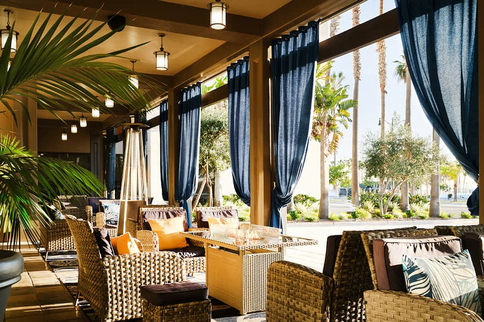 Covered patio seating area with woven furniture, blue curtains, wooden beams, and palm tree views
