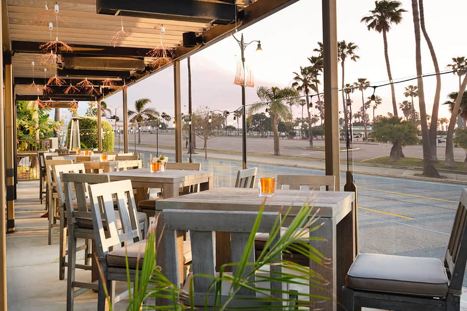 Modern outdoor restaurant with pergola, wooden tables, palm trees visible, and tropical sunset ambiance