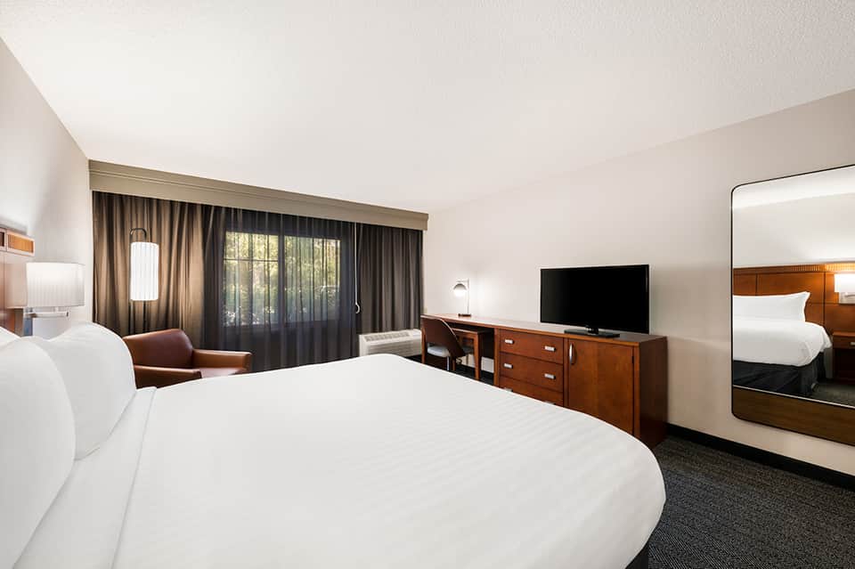 Modern hotel room with white bedding, wooden furniture, dark curtains, and flat-screen TV