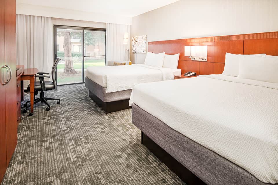 Hotel guest room with two queen beds, orange accent wall, desk, sliding glass doors to lawn, and modern furnishings