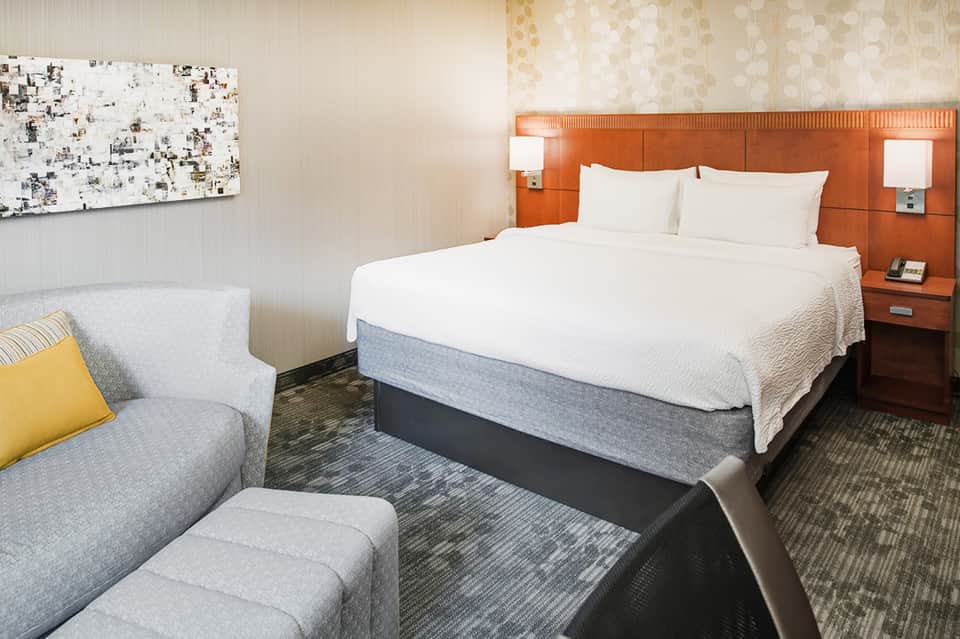 Hotel room with queen bed, orange headboard, white linens, and lounge seating area with contemporary decor