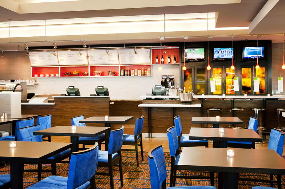 Modern hotel cafe with blue chairs, dark tables, counter service, and TV screens