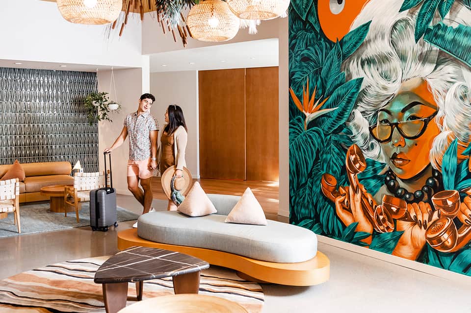 Modern hotel lobby with tropical mural art, woven pendant lights, and guests with luggage