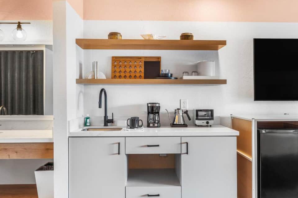 Modern hotel kitchenette with white cabinetry, wooden shelves, black sink fixture, and stainless appliances