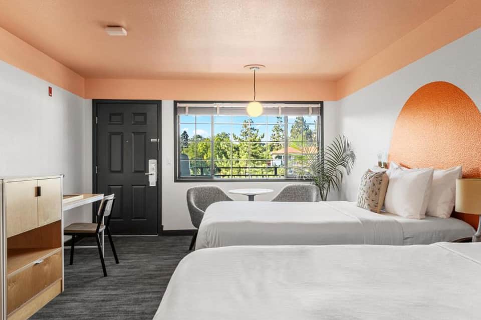 Contemporary hotel room with two beds, orange accent wall, desk, and large windows with views
