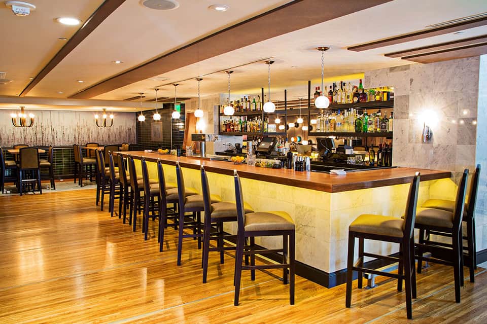 Upscale hotel bar with yellow-lit counter, wooden furnishings, pendant lights, and full liquor shelves