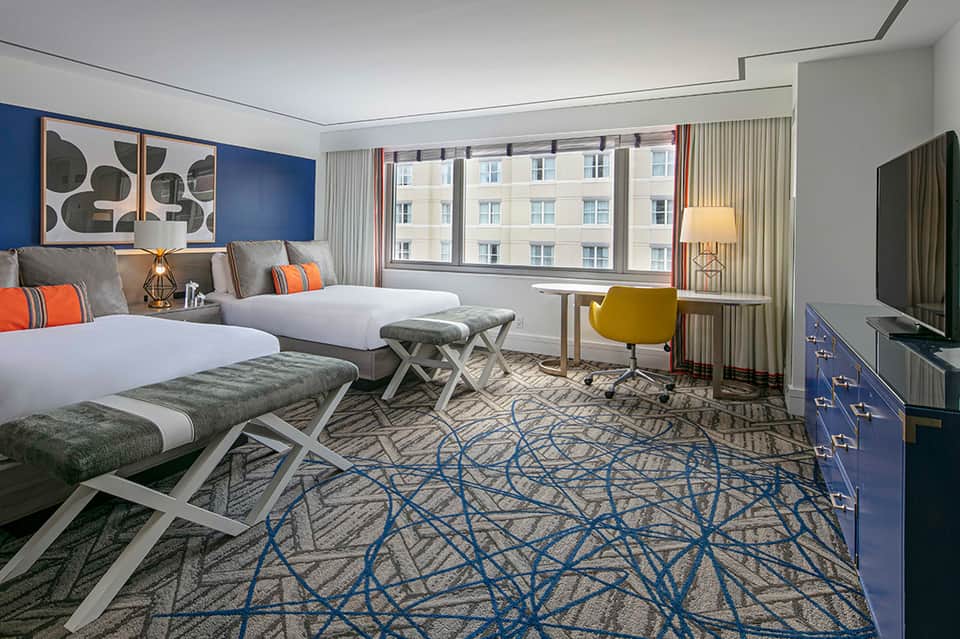 Contemporary hotel room with two beds, blue accent wall, orange pillows, and city views