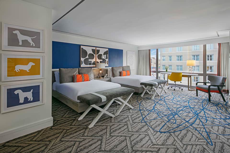 Modern hotel bedroom with blue accent wall, orange pillows, and city views
