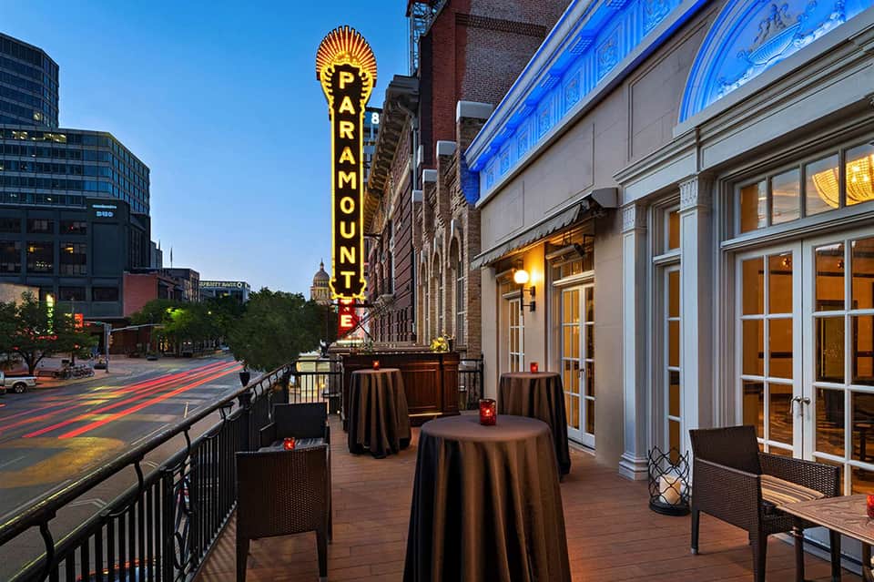 Hotel rooftop terrace with Paramount theater neon sign, dark round tables, and urban skyline at dusk