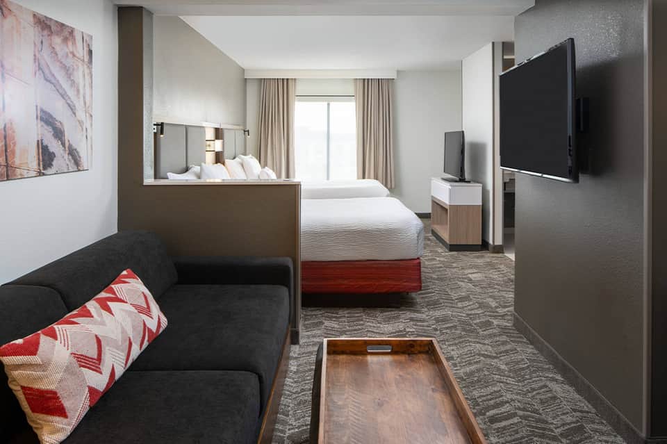 Modern hotel room with queen beds, dark sofa, TV, and neutral contemporary furnishings